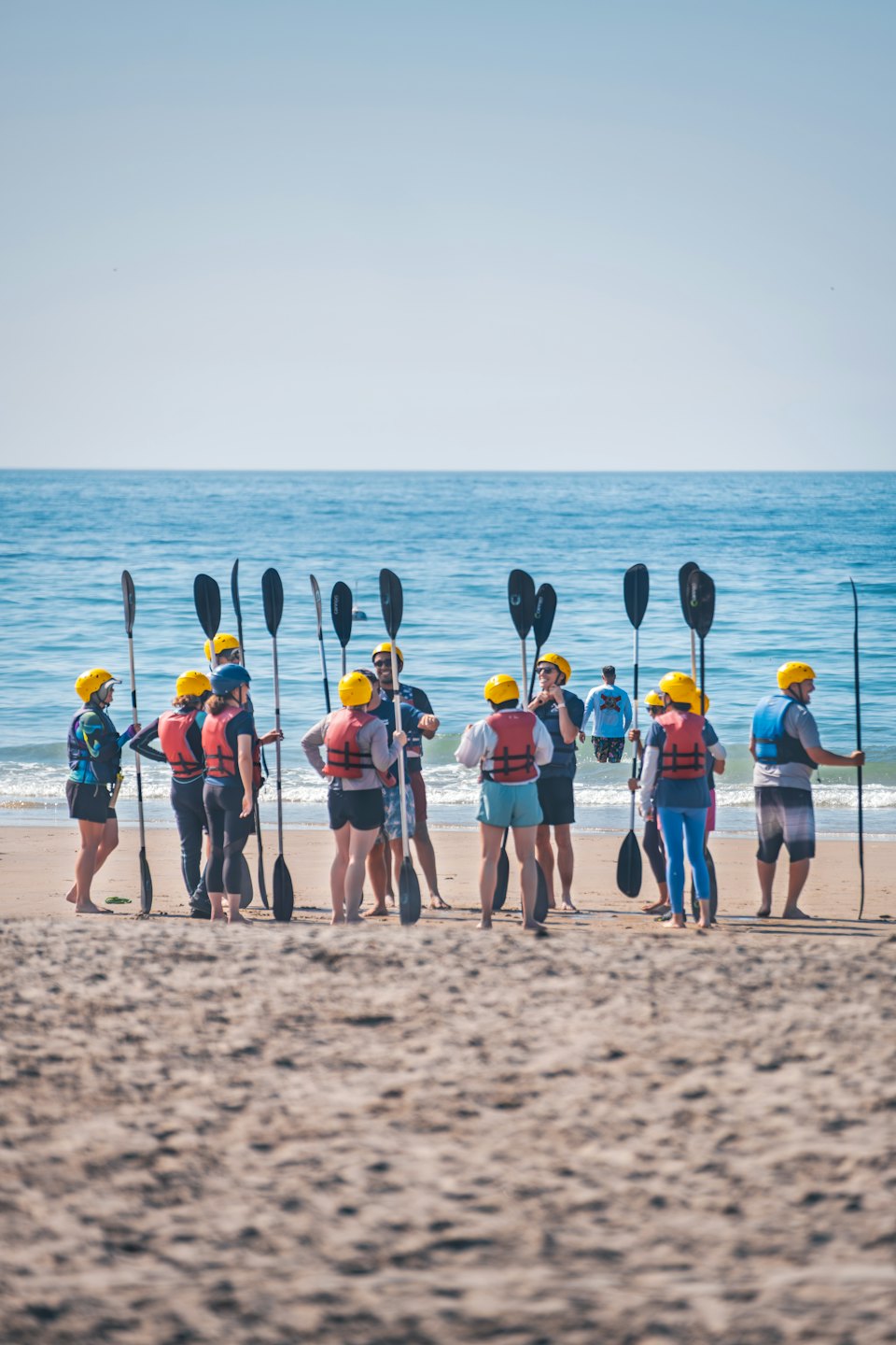 Persone con giubbotti di salvataggio e caschi gialli che tengono pagaie su una spiaggia.