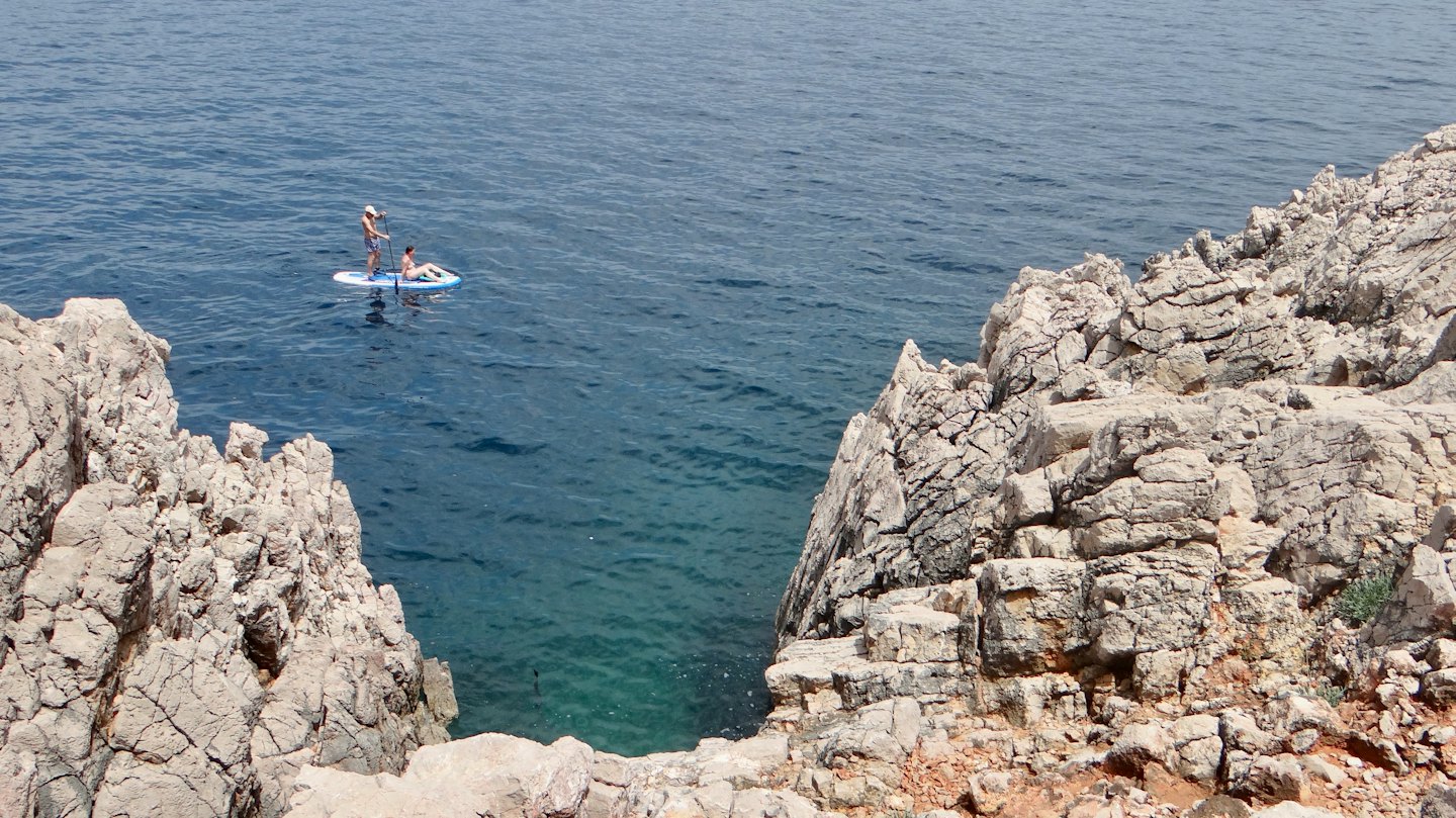 Due persone in piedi su tavole da paddleboard su un mare blu-verde limpido vicino a scogliere rocciose.